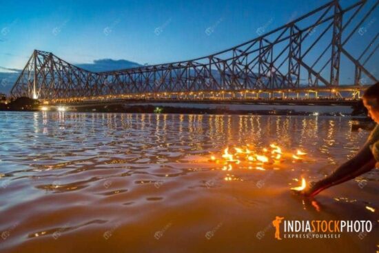 Old woman floats clay diya lamps in the Ganges with view of Howrah bridge at Kolkata