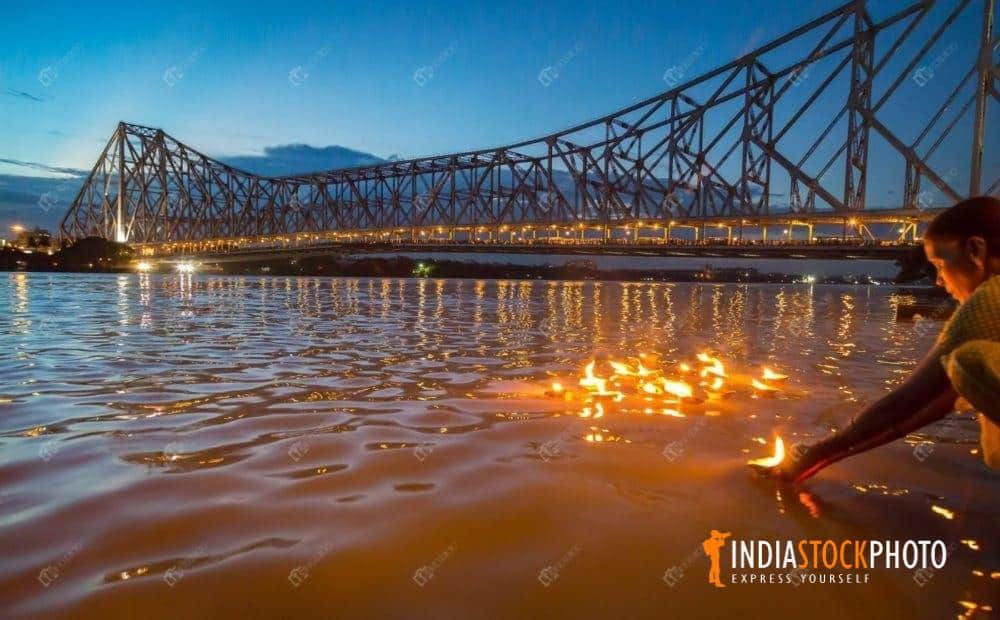 Old woman floats clay diya lamps in the Ganges with view of Howrah bridge at Kolkata